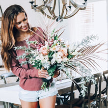 Load image into Gallery viewer, Smiling woman holding unique flower bouquet, modern dining room and chandelier in background.
